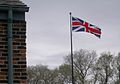Flag on pole before grey sky and trees in early spring. A modern brick building is visible on the left.