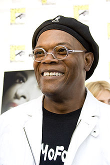 An African-American man is at the centre of the image looking to the left and smiling. He is wearing a hat, glasses, a white jacket and a black t-shirt that says "MoFo".